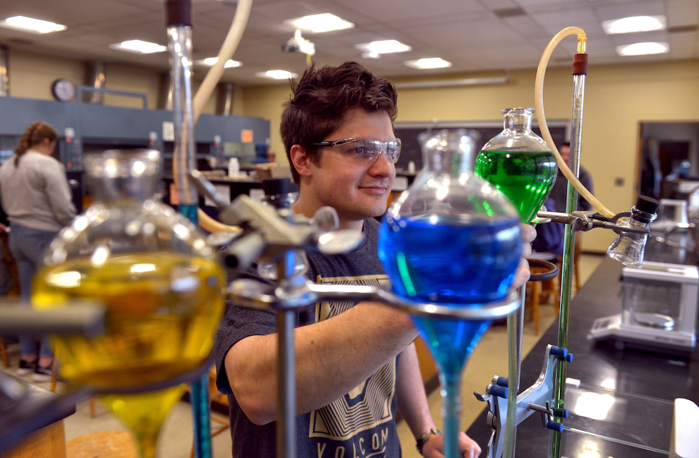 A chemistry student smiles while examining glass containers of colored liquid